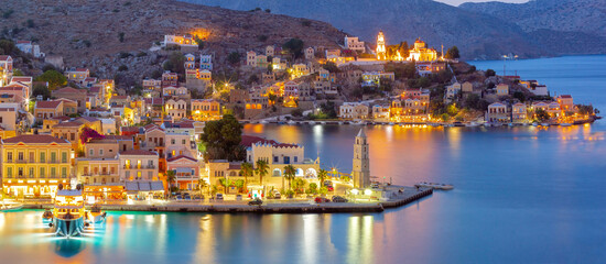 Fototapeta premium Panoramic view of Symi Harbor at night with illuminated buildings and boats, Greece