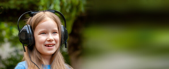 One single happy cheerful school age child, girl wearing large headphones listening to music outdoors, simple portrait, face up close, wide shot blurry background, shallow DOF, copy space, web banner