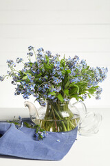Fototapeta premium Bouquet of beautiful forget-me-not flowers in glass teapot and blue cloth on white table