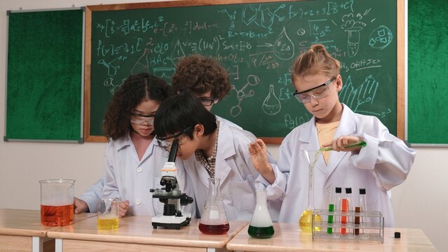 Smart caucasian teacher looking under microscope while diverse children doing experiment at STEM science lesson. Professional happy scientist inspect biochemical example by using microscope. Pedagogy.