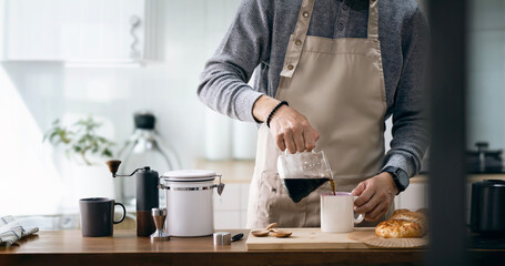 Asian man in apron pouring coffee in cup while standing near cooking surface in kitchen - Powered by Adobe