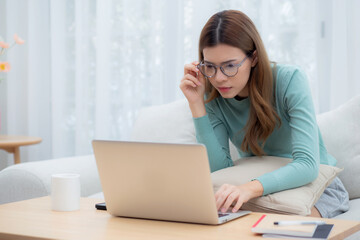 Concentrated young asian woman wearing glasses working on laptop with commitment and ambition sitting on sofa in living room at home, work from home, freelance working online, business and lifestyle.