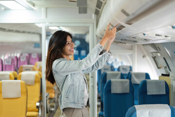 Young Woman Storing Luggage in Overhead Compartment on Airplane, Smiling Passenger in Modern...