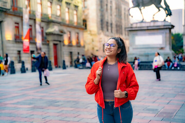 Smiling young Latin woman with long hair and eyeglasses in Munal Museum in Mexico City Downtown smiling and zipping up red jacket against gray door