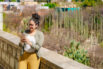 A young Latina, smiling and engaged, texting with a view of the Ethnobotanical Garden from Santo...