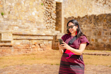 Joyful young Latina woman in traditional dress takes a selfie at Monte Alban ruins in Oaxaca, Mexico, capturing her adventure with smartphone.