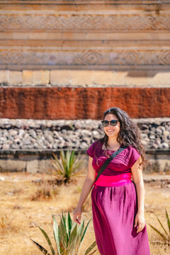 Young Latina woman smiling at Mitla ruins, Oaxaca, Mexico, wearing traditional dress and sunglasses on a sunny day.