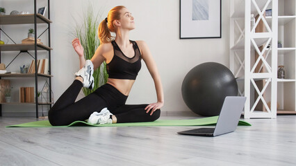 Stretch exercise. Online session. Gymnastic class. Muscle soreness relief. Free movement. Female fitness instructor in sportswear pull leg on yoga mat with laptop at home interior.