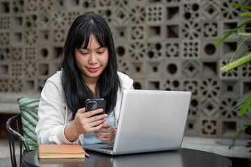 A young Asian woman is reading messages on her smartphone while working remotely from a coffee shop.