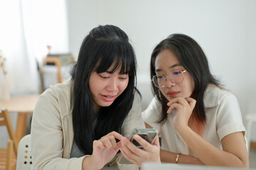 Two Asian female friends are watching something on a smartphone together while sitting in a cafe.