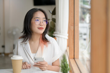 A young, positive Asian woman is daydreaming while gazing at the view outside of a coffee shop.