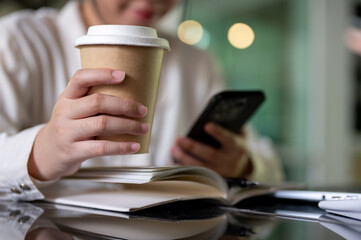 A cropped image of a businesswoman holding a coffee cup while using her smartphone at her desk.