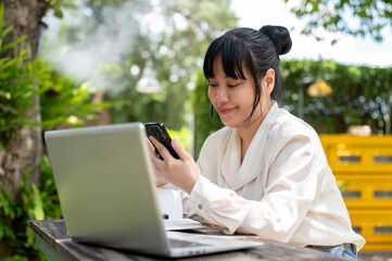 A positive Asian woman using her smartphone, reading messages while sitting at a table in a garden.