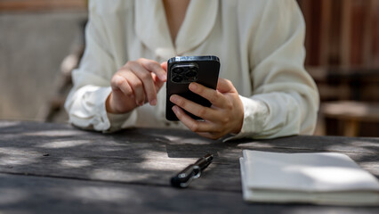 A cropped image of a woman using her smartphone at a wooden table in a garden on a bright day.