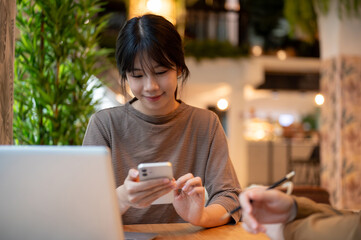 Beautiful Asian woman is reading messages on her smartphone while working remotely in a coffee shop.
