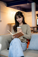 A pretty, happy Asian woman reading a book and sipping coffee while sitting in a cozy coffee shop.