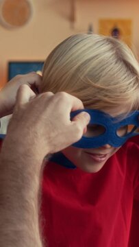 Vertical over shoulder of dad helping tween son to wear blue superhero eye mask while having fun together in apartment