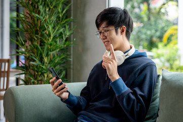 A young Asian man is talking on a video call on his phone with his friend while relaxing at home. - Powered by Adobe