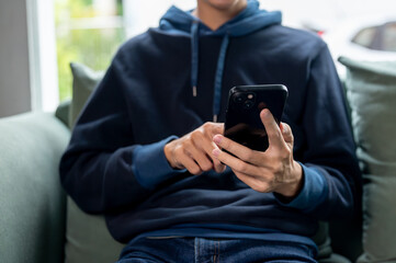 A cropped image of a young man in a blue hoodie using his smartphone on a couch at home.