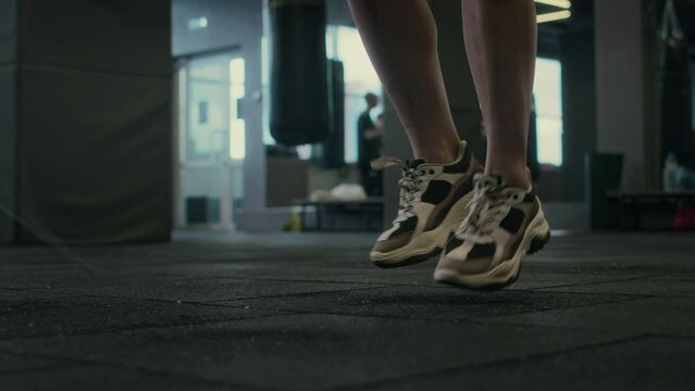 Close-up shot of feet of unrecognizable young female sportswoman in trainers walking up over rubber floor tiles towards camera and beginning to warm up with jumping rope for intense cardio workout