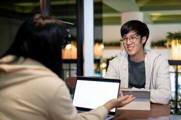 Obraz premium A happy Asian male student is talking with his friend while doing homework together in a cafe.