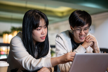 Fototapeta premium A smart Asian female student is helping her friend with a project, explaining something on a laptop.