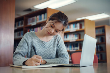 Asian woman student writing notes with textbooks for knowledge, information and literature project in library for research, studying and learning.
