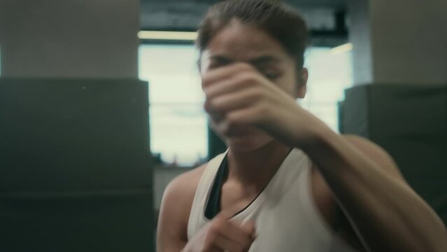 Medium close-up handheld POV shot of muscular young female athlete shadowboxing energetically at gym, working on punch, jab, lunge maneuvers while facing towards camera