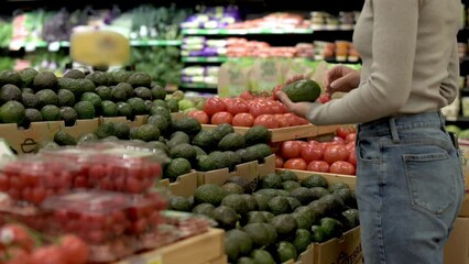 A woman in a beige sweater and blue jeans picking avocados from a display in the produce section of a grocery store, with tomatoes and other fresh produce in the background.