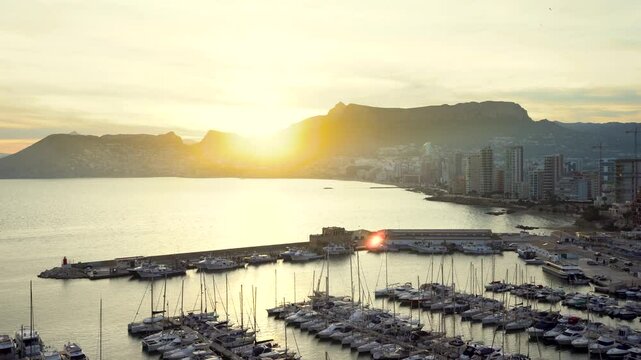 Panoramic high angle view of Marina of Calpe and part of city. Sun settles down behind the mountains. Skyscrapers in first line of the beach. Camera panning right. Travel destination.