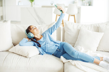 Little kid girl sitting on sofa, using smartphone with headphones, take selfie. Kids and gadgets...