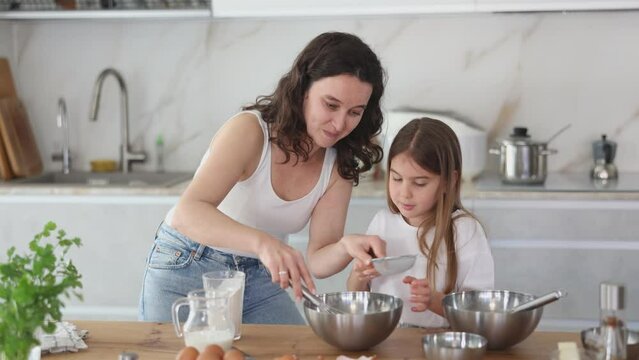 Happy mother and her daughter adding flour in bowl and whisking ingredients using metal corolla in the kitchen