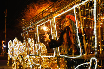 Woman holding sparkler night while celebrating Christmas outside. Dressed in a fur coat and a red headband. Blurred christmas decorations in the background. Selective focus