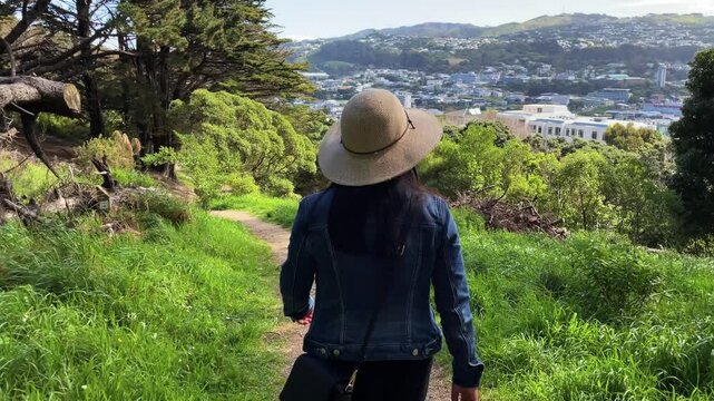 A woman with black hair wearing a sun hat and blue denim jacket walks down a dirt hiking trail through a forest towards the view of the city of Wellington, New Zealand