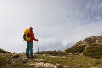 A person wearing a red jacket and blue pants is standing on a hillside with a yellow backpack