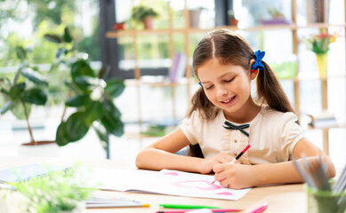 Portrait of pretty little schoolgirl sitting at table desk, drawing red heart, ready to study. Happy smiling cute school girl learn in classroom, indoors. Primary education concept image.