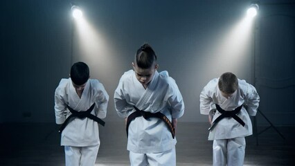 Three children wearing karate uniforms stand confidently in front of bright spotlight, showcasing their martial arts skills with determination and focus. Boys training karate at smoky background - Powered by Adobe