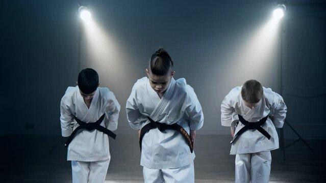 Three children wearing karate uniforms stand confidently in front of bright spotlight, showcasing their martial arts skills with determination and focus. Boys training karate at smoky background