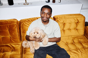 Minimal portrait of smiling African American man holding little dog sitting on couch and looking at...