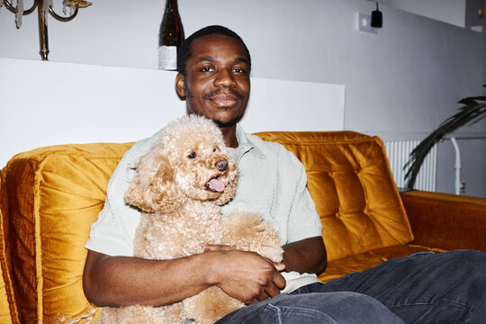 Minimal portrait of African American man with cute dog in lap sitting on couch and looking at camera, with flash