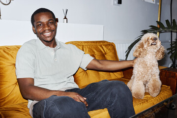Minimal portrait of young African American man sitting on velvet couch with cute dog and smiling happily at camera, copy space