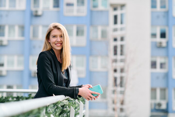 Fototapeta premium Blonde Businesswoman in Black Suit Using Smartphone on Break in Modern Urban Environment.