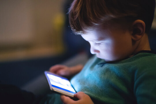 Cute boy sitting on bed watching something on smartphone. Children's screen time.