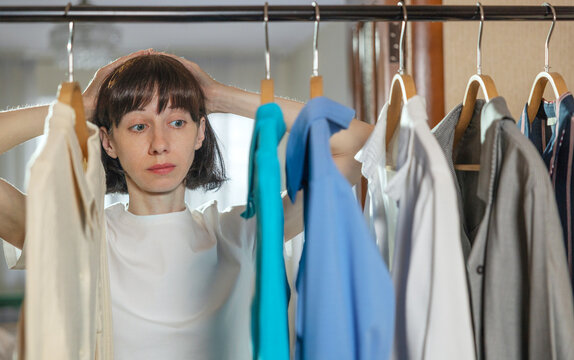 Confused mature woman choosing clothes hanged on rack at home