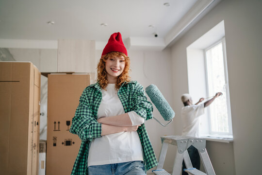 Happy young woman standing with paint roller near friend working in background