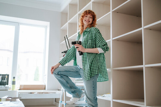 Happy woman standing with drill on step ladder near cabinet at home