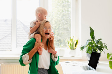 Cheerful mother carrying son on shoulders and working at home