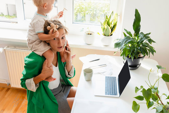 Freelancer talking on smart phone and carrying son over shoulders at desk