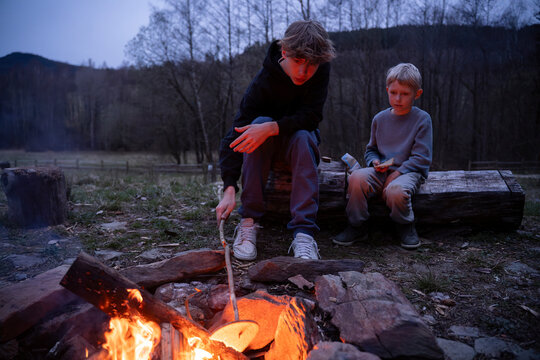 Teenage boy sitting with brother near bonfire in back yard at dusk