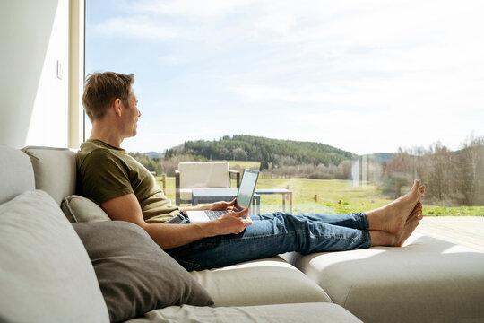 Man with laptop looking at view through large window sitting on sofa in living room